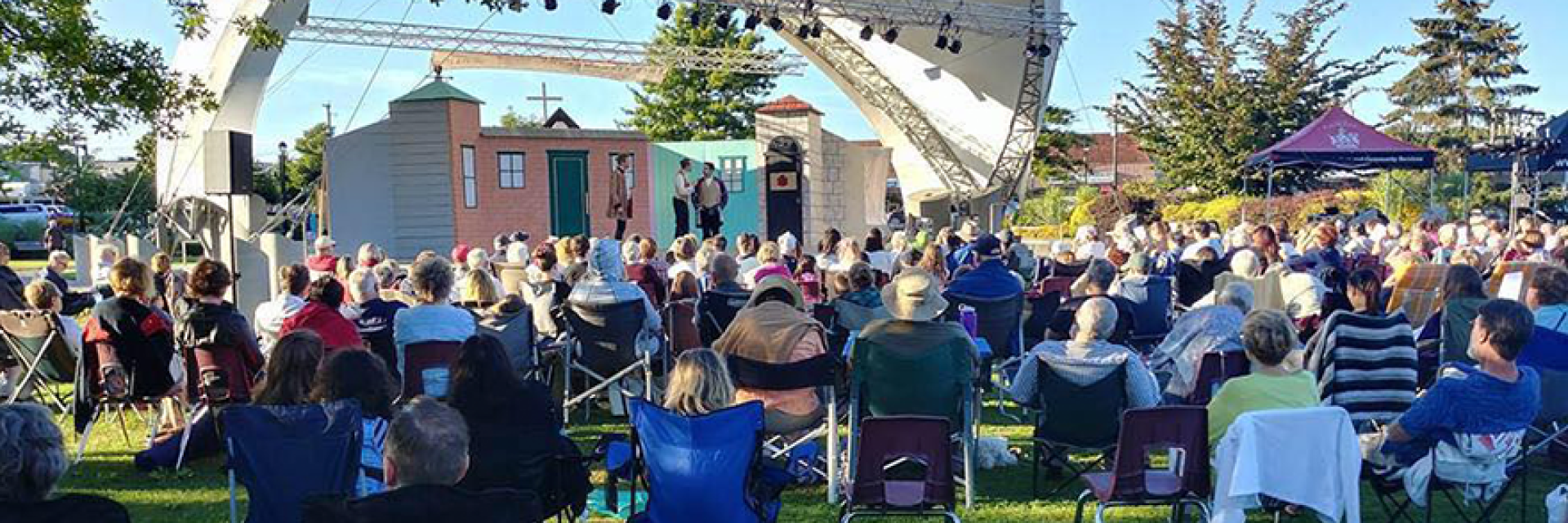 Audience sitting outdoors on lawn chairs watching a live theatre performance.