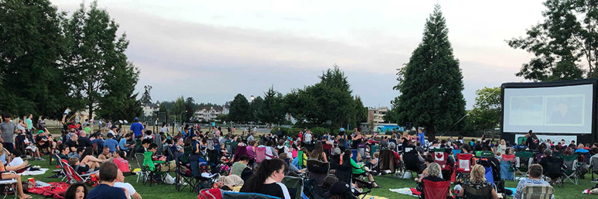Crowd of families sitting on lawn chairs and blankets in the park getting ready to watch a movie outdoors on an inflatable screen.