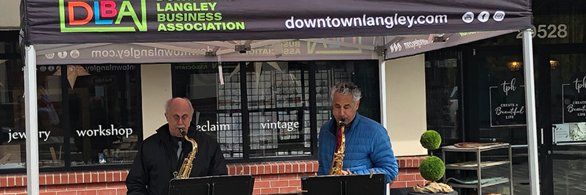 Two men playing the saxophone outdoors under a pop up tent.