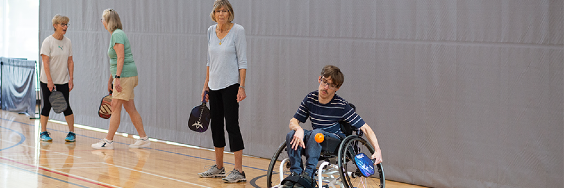Four participants, three female and one male, participants in gymnasium pickleball; one participant is in a wheelchair.