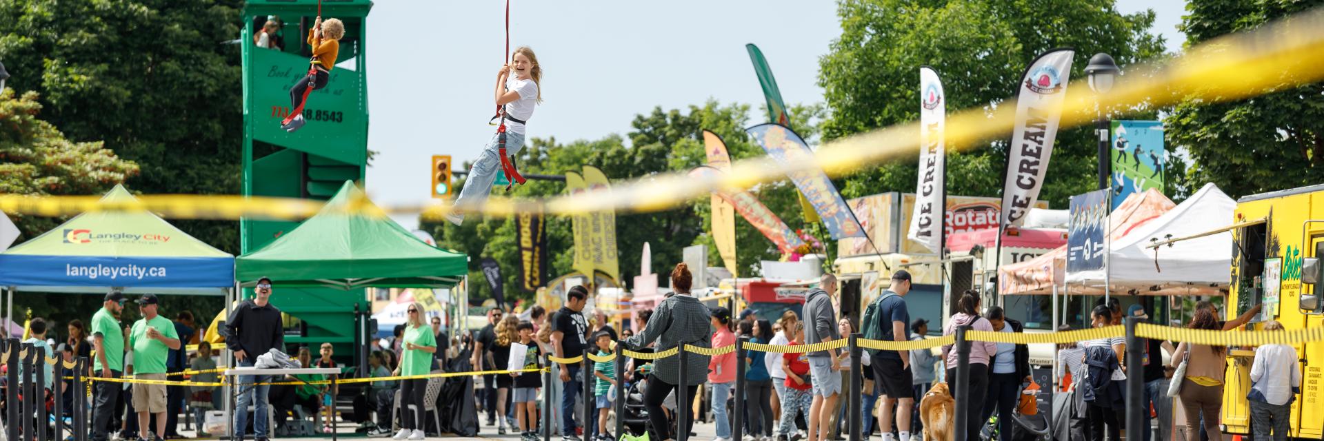 Two people on a zipline at a community festival, surrounded by other people walking through event space and visiting food trucks.