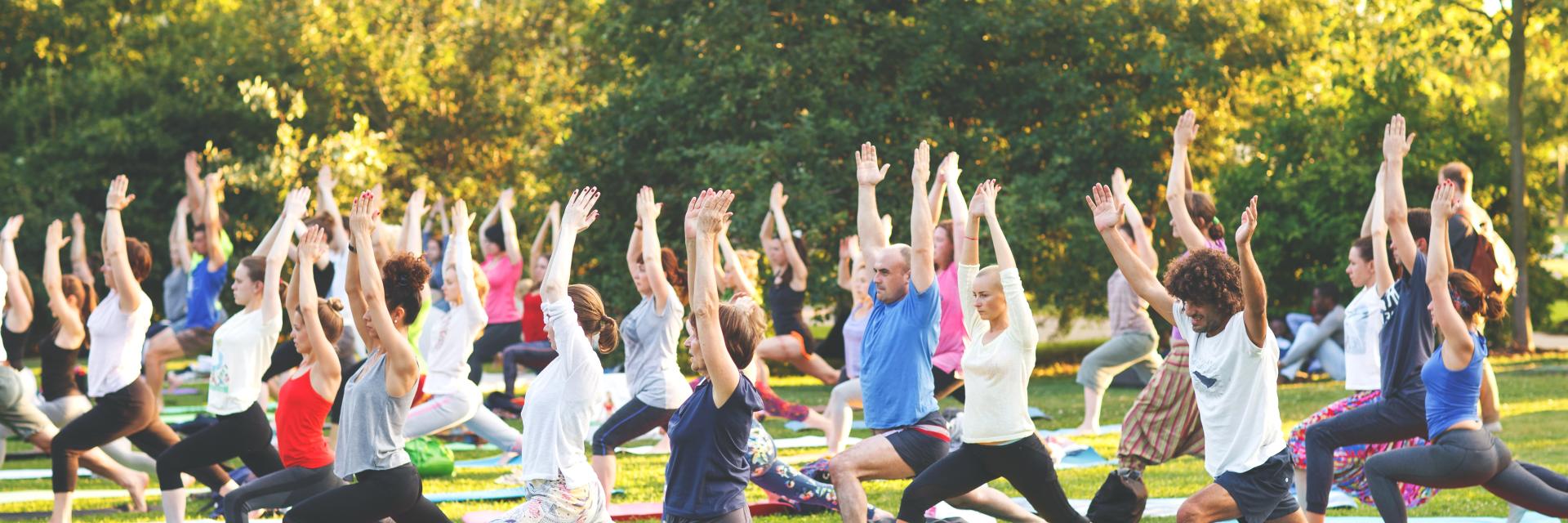 Multiple people of all genders, ages, and ethnicity doing yoga exercise outdoors in a park.