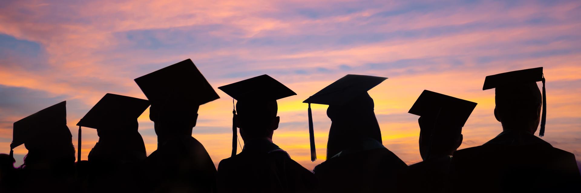 Silhouettes of people wearing graduation hats standing with a sunset in the background.