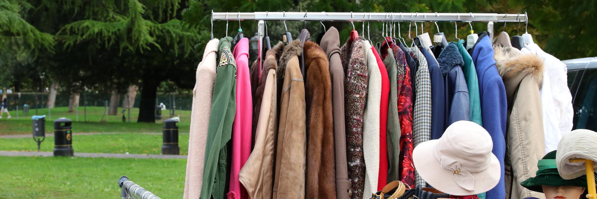 Rack and tables with clothing and other wearable items for sale at an outdoor garage sale located in a park.