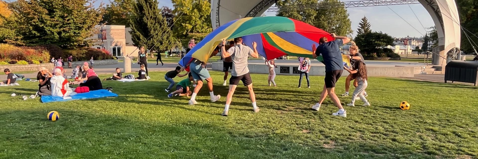 Group of people, adults and kids, playing outside in park with a large play parachute.