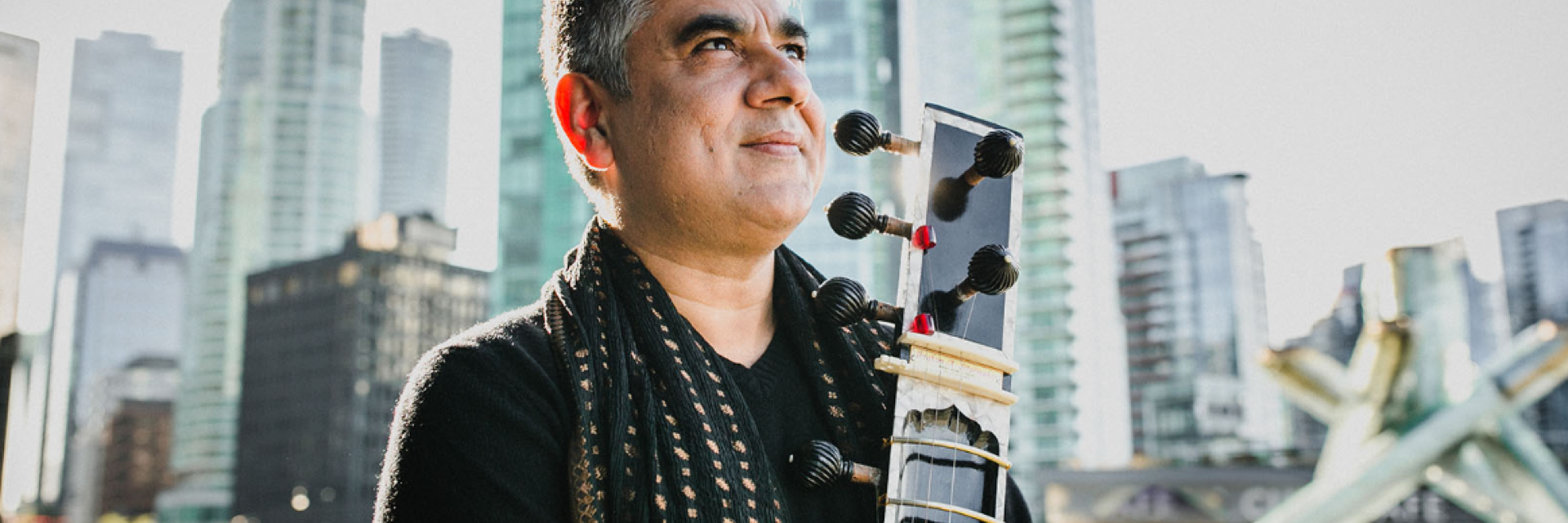 Male musician standing holding instrument outdoors with a city scape background.