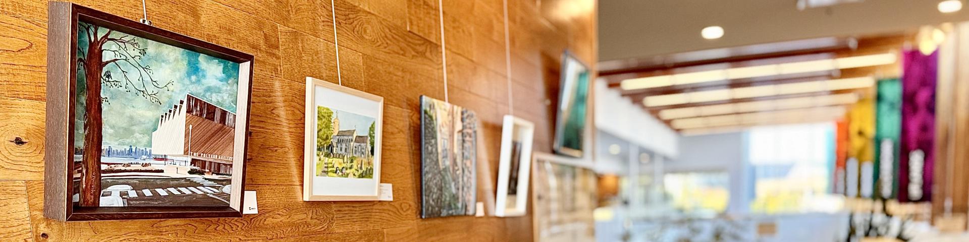 Five framed artworks displayed in A Step Above Cultural Gallery, hung on a horizontal wooden panel wall using a wired hanging system in a gallery setting.