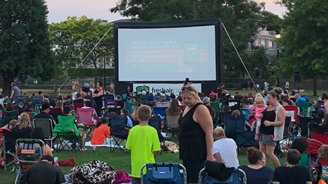 Crowd of all ages sitting on lawn chairs waiting to watch a movie on an inflatable screen in a park.