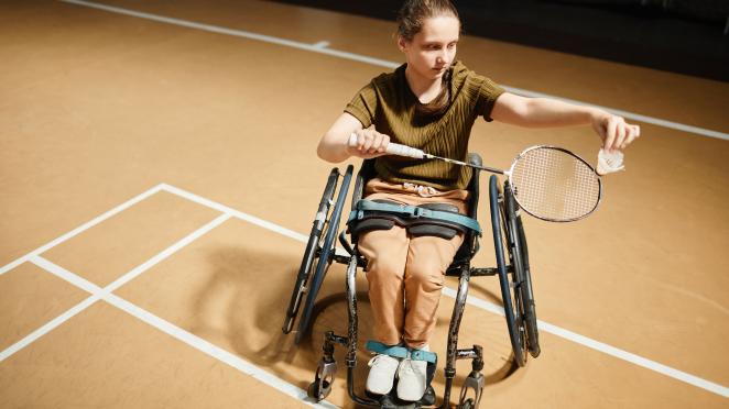 Individual in a wheelchair playing badminton inside a gymnasium.