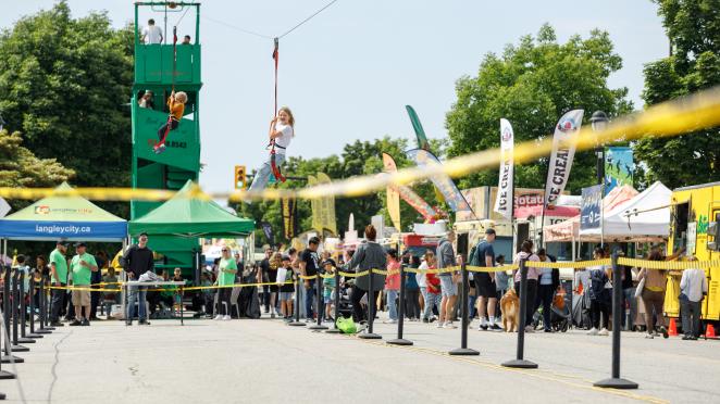 Two people on a zipline at a community festival, surrounded by other people walking through event space and visiting food trucks.