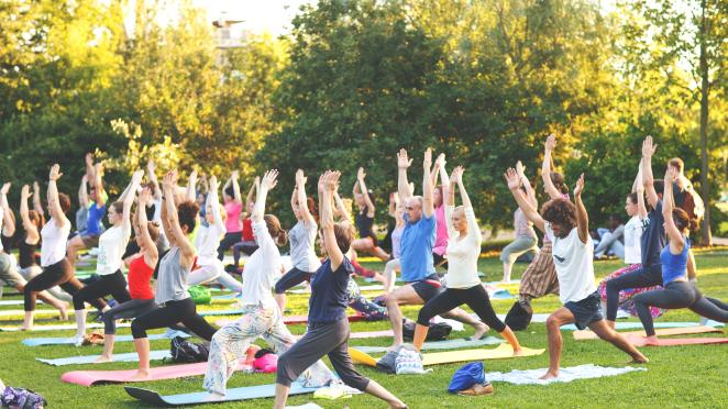 Multiple people of all genders, ages, and ethnicity doing yoga exercise outdoors in a park.