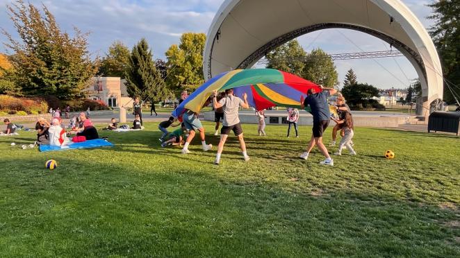 Group of people, adults and kids, playing outside in park with a large play parachute.