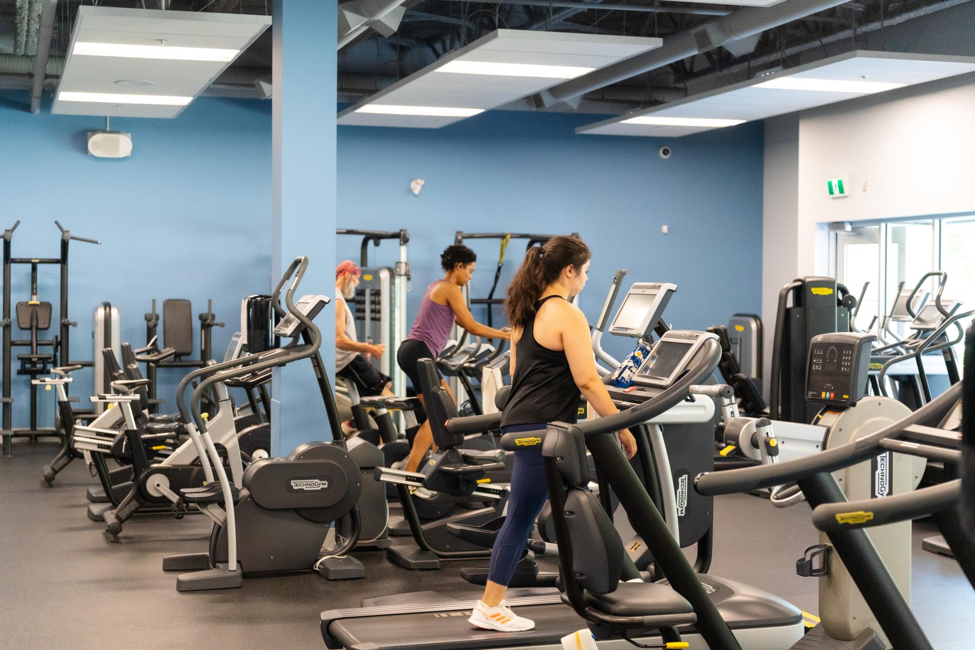 Three adults working out in a weight room; one person on a treadmill, one person on a elliptical, and one person on a spin bike.